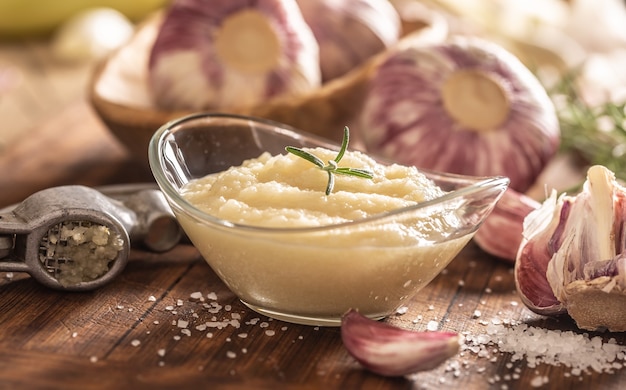 Foto pasta de alho em uma tigela de vidro com alho descascado, sal e cabeças de alho.