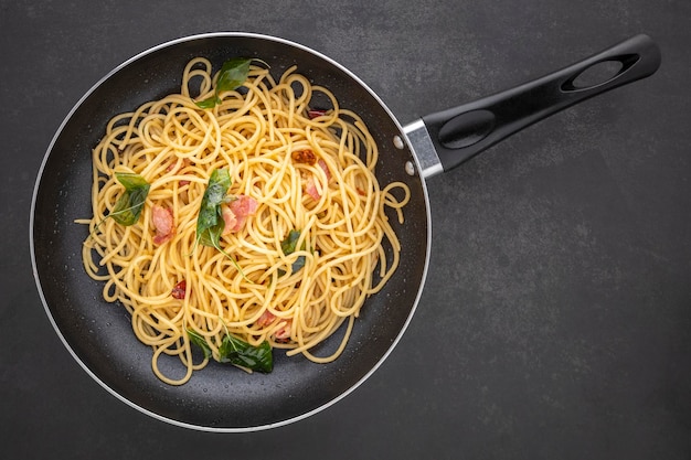 Foto pasta de espaguete com alho picante seco, manjericão doce e bacon em panela preta, vista de cima