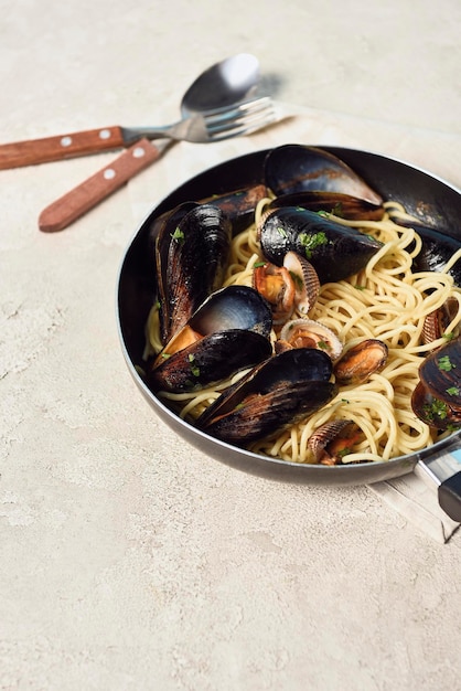 Foto pasta deliciosa con mariscos en la sartén cerca de los cubiertos sobre un fondo gris texturizado