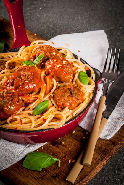 Pasta de espagueti con albóndigas, salsa de tomate y albahaca en sartén de hierro fundido rojo, sobre mesa de piedra negra con tabla para cortar