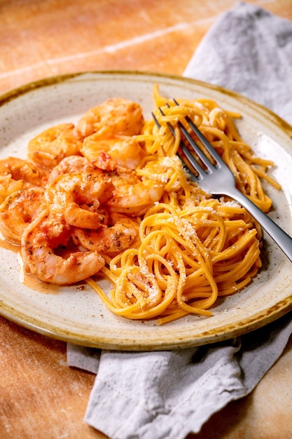 Pasta de espagueti de tomate con camarones, gambas en salsa y queso parmesano servido en plato de cerámica manchado con tenedor y servilleta de tela sobre muro de piedra marrón.