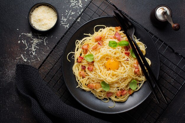 Pasta espaguetis carbonara con tocino, queso parmesano, yema de huevo y hojas de albahaca sobre superficie negra. Plato tradicional italiano