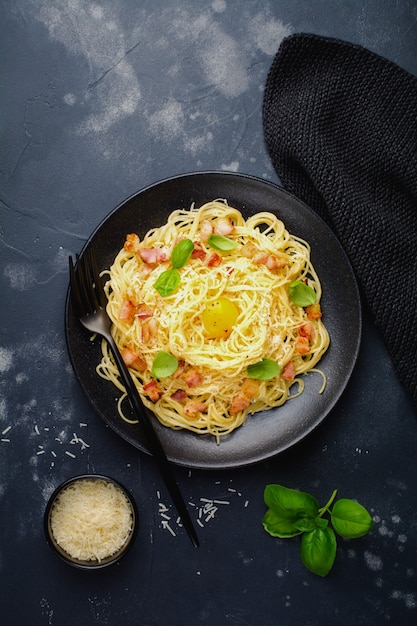 Pasta espaguetis carbonara con tocino, queso parmesano, yema de huevo y hojas de albahaca sobre superficie negra. Plato tradicional italiano