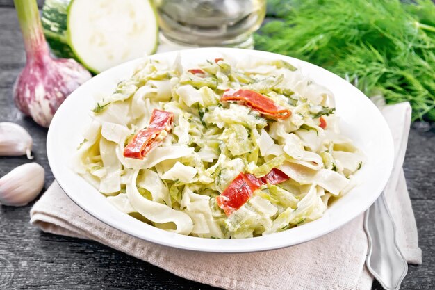 Pasta fettuccine con calabacín y pimiento rojo picante en salsa cremosa en un plato blanco sobre una toalla de cocina, ajo y un tenedor sobre fondo de madera negra