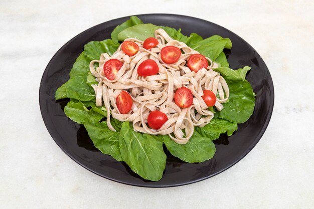 Pasta integral con hojas verdes de rúcula y tomate cherry sobre placa negra y mármol blanco