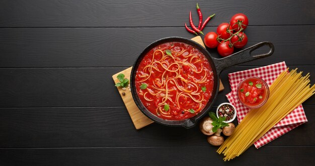 Pasta italiana hirviendo casera con salsa de tomate en una sartén de hierro fundido servida con ají rojo, albahaca fresca, tomates cherry y especias sobre una mesa de madera rústica negra, concepto de comida para cocinar