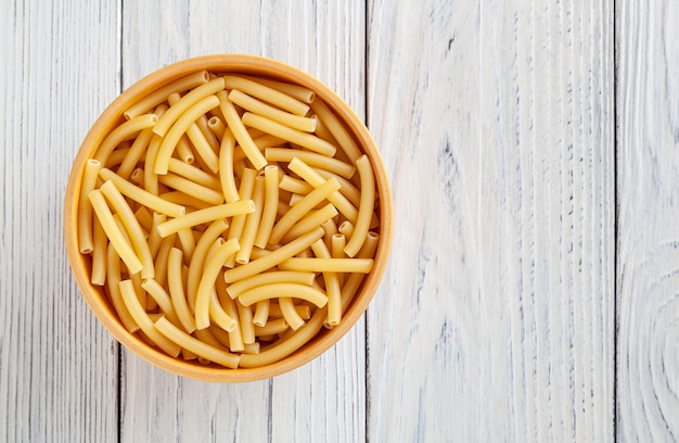 Pasta macarrones sin cocer en un tazón de madera sobre fondo blanco de madera