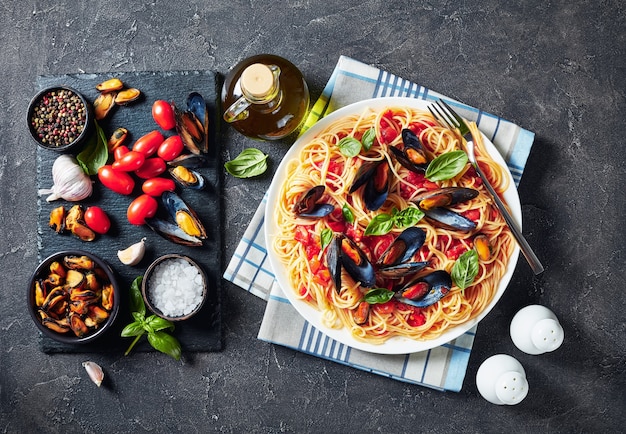 Pasta con mariscos en salsa de tomate picante en un plato blanco sobre una mesa de hormigón con ingredientes en una bandeja de pizarra negra, vista horizontal, primer plano