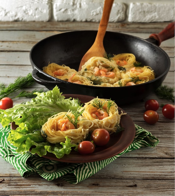 Pasta mit Garnelen in Form von Nestern. Eine Portion Nudeln auf einem Teller mit Salat. In der Nähe von Pfanne mit Nudeln, Tomaten und Dill