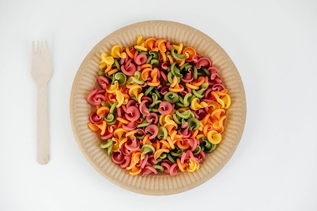 Pasta multicolor en un plato de papel desechable con un tenedor de madera sobre un fondo blanco.