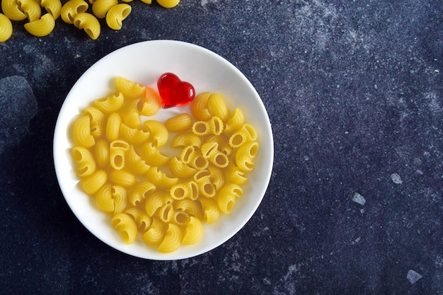 Pasta no cocida en un plato blanco con un corazón rojo sobre un fondo oscuro con espacio para copiar