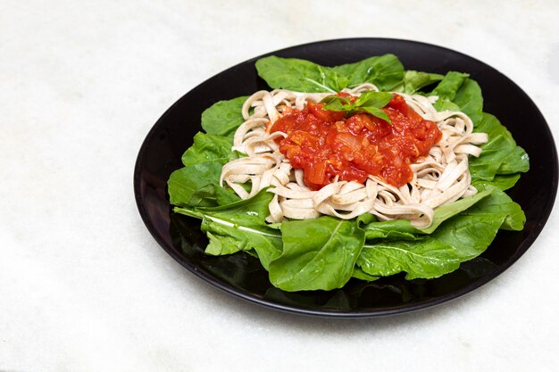 Pasta de pasta integral con hojas verdes de rúcula, salsa roja y vinagre balsámico sobre placa negra y fondo de mármol blanco