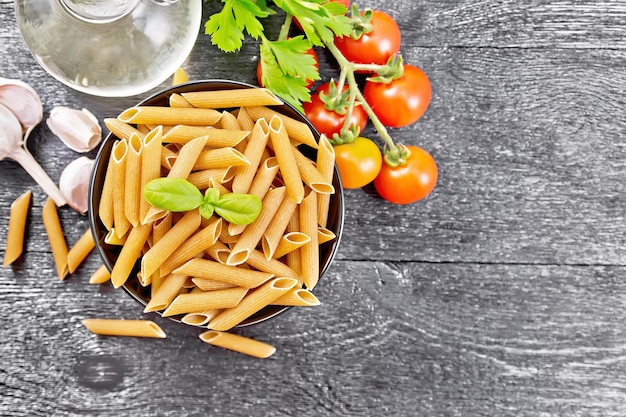 Pasta penne de harina integral en un bol, tomates, ajo, aceite vegetal en un decantador y perejil sobre fondo de tablero de madera desde arriba