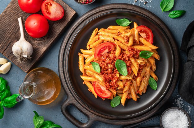 Pasta penne con salsa boloñesa en un plato negro sobre un fondo de ajo, tomate y albahaca. Vista superior, primer plano.