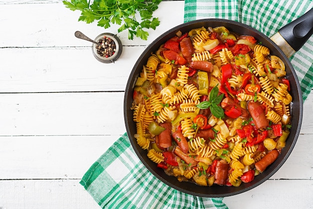 Pasta con salsa de tomate con salchicha, tomates, albahaca verde decorado en una sartén sobre un fondo de madera blanca.