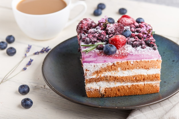 Pastel de bayas con crema de leche y mermelada de arándanos en placa de cerámica azul con taza de café y arándanos frescos sobre una superficie de madera blanca. Vista lateral, enfoque selectivo.