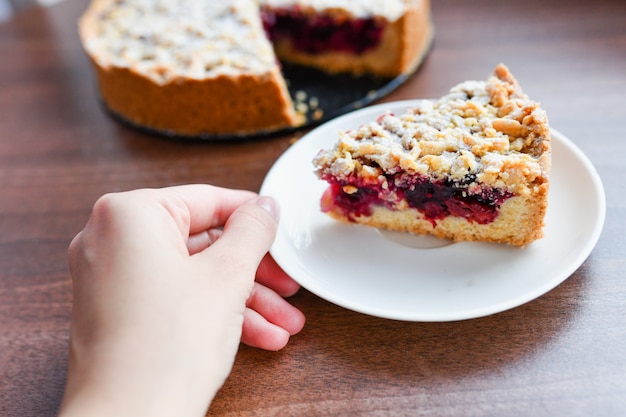 Pastel con bayas: frambuesas, fresas, grosellas, en un plato blanco, la mano de una mujer sosteniendo un pedazo de pastel en una espátula. Sobre un fondo de madera, en el fondo servilleta de lino