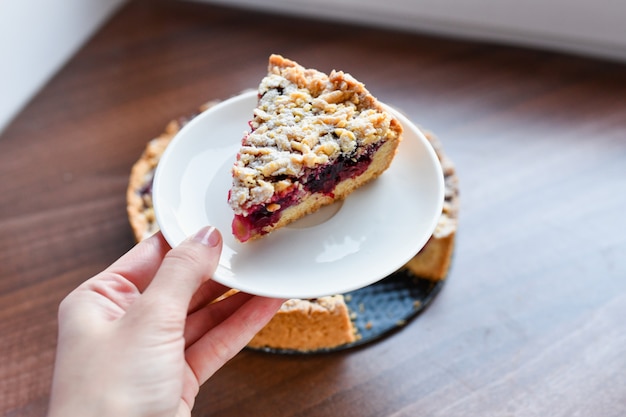 Pastel con bayas: frambuesas, fresas, grosellas, en un plato blanco, la mano de una mujer sosteniendo un pedazo de pastel en una espátula. Sobre un fondo de madera, en el fondo servilleta de lino