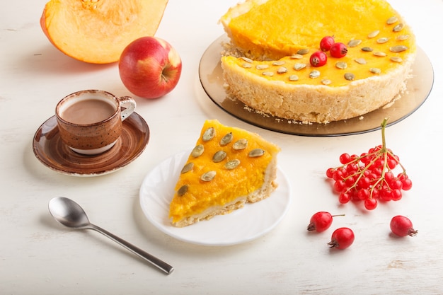 Pastel de calabaza dulce americano tradicional decorado con bayas rojas de espino y semillas de calabaza con una taza de café sobre un fondo blanco de madera. vista lateral.