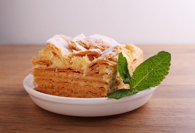 Foto pastel de capas napoleón con una rodaja de crema de leche de vainilla decorada con menta, en un plato sobre un fondo de madera.