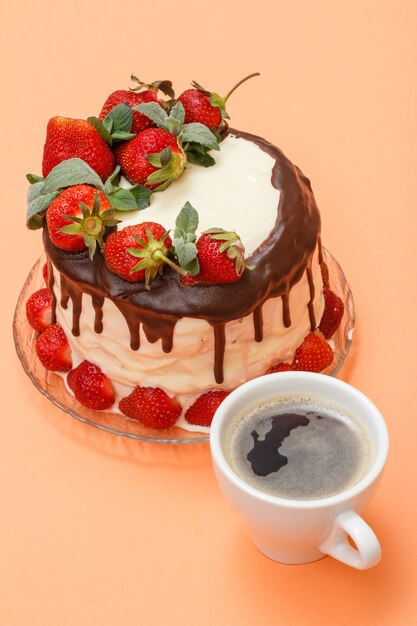 Pastel de chocolate casero decorado con fresas frescas y hojas de menta en un plato de vidrio y una taza de café sobre fondo de color melocotón