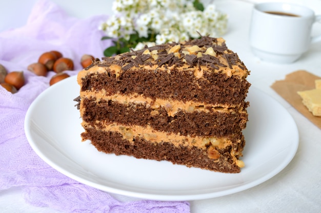 Pastel de chocolate con crema de nuez sobre una mesa de madera blanca. Un pedazo de pastel en un plato y una taza de café.