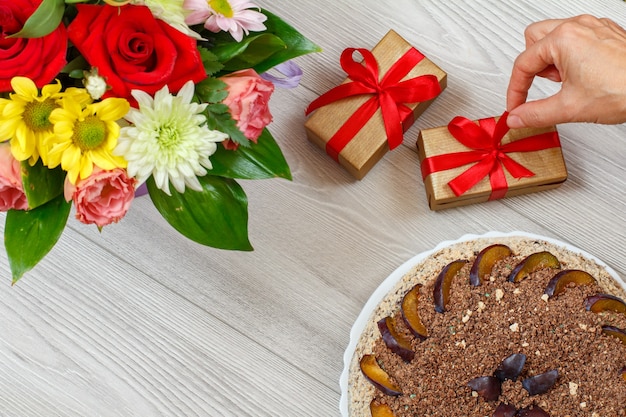 Pastel de chocolate decorado con ciruelas, un ramo de flores y cajas de regalo sobre las tablas de madera gris. Vista superior.