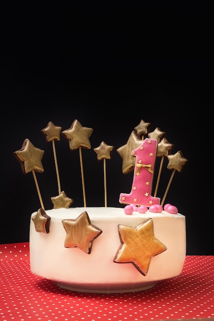 Pastel de cumpleaños decorado con rosa número 1 y estrellas doradas de pan de jengibre en una pared oscura.