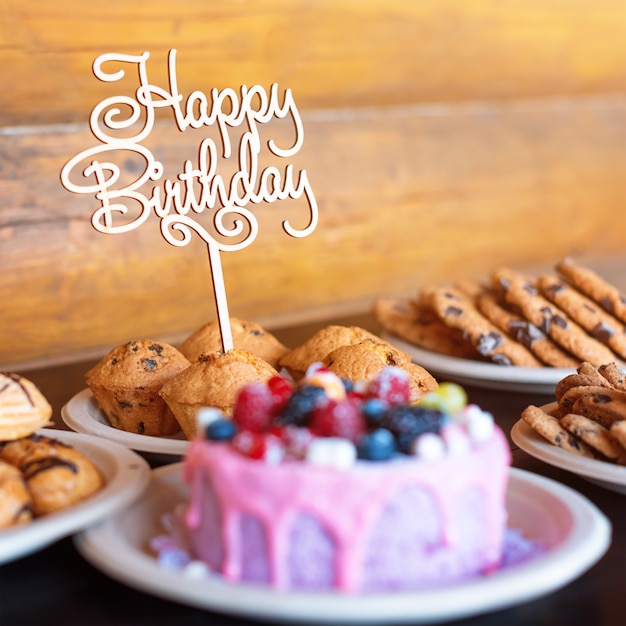 Pastel de cumpleaños y magdalenas con cartel de saludo de madera en la pared rústica. Madera canta con letras feliz cumpleaños y dulces de vacaciones.