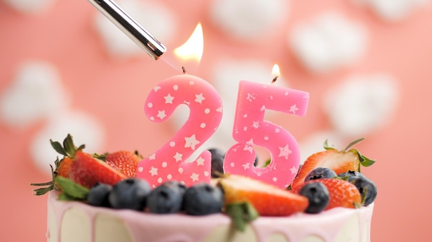 Pastel de cumpleaños número 25 vela rosa en un hermoso pastel con bayas y encendedor con fuego contra el fondo de nubes blancas y cielo rosa Vista de cerca