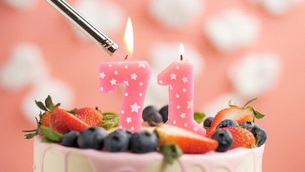 Pastel de cumpleaños número 71 vela rosa en un hermoso pastel con bayas y encendedor con fuego contra el fondo de nubes blancas y cielo rosa Vista de cerca