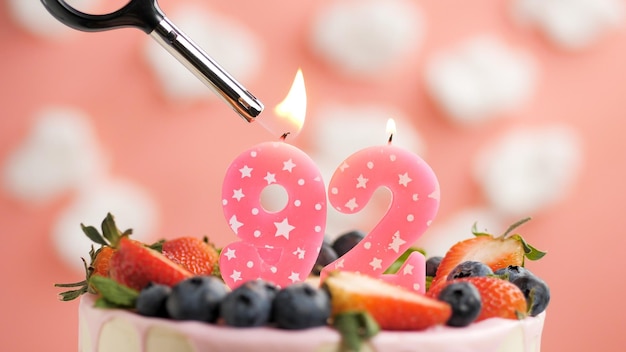 Pastel de cumpleaños número 92 vela rosa en un hermoso pastel con bayas y encendedor con fuego contra el fondo de nubes blancas y cielo rosa Vista de cerca