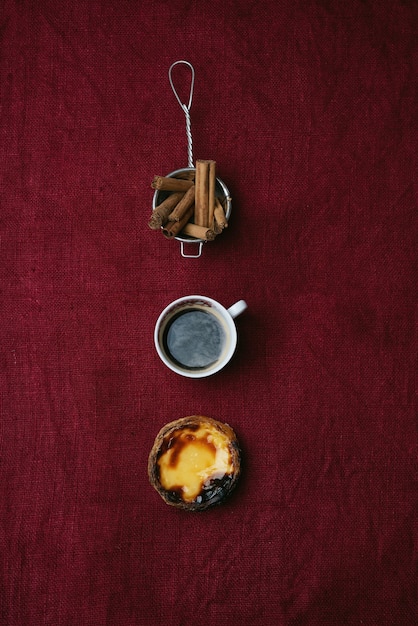 Pastel de nata. Sobremesa tradicional portuguesa, torta de ovo, xícara de café e pau de canela na peneira sobre fundo de matéria têxtil. Vista do topo
