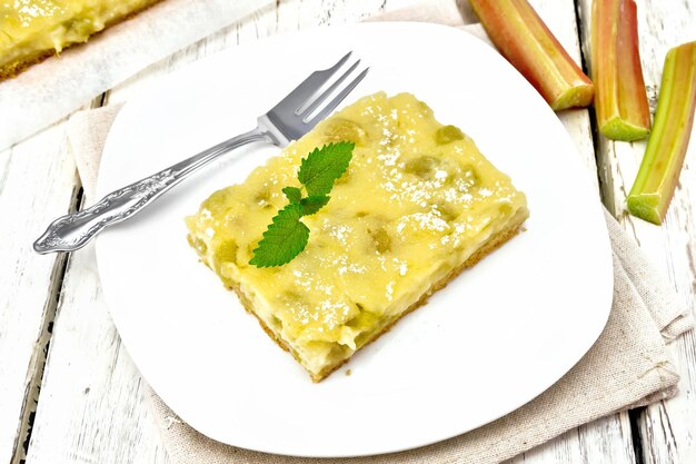 Pastel dulce con relleno de pudín de vainilla, ruibarbo y crema en un plato con menta en una servilleta con el telón de fondo de la tabla de madera clara