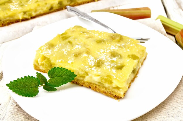 Pastel dulce con relleno de pudín de vainilla, ruibarbo y crema en un plato con menta sobre una toalla de cocina con el telón de fondo de la tabla de madera