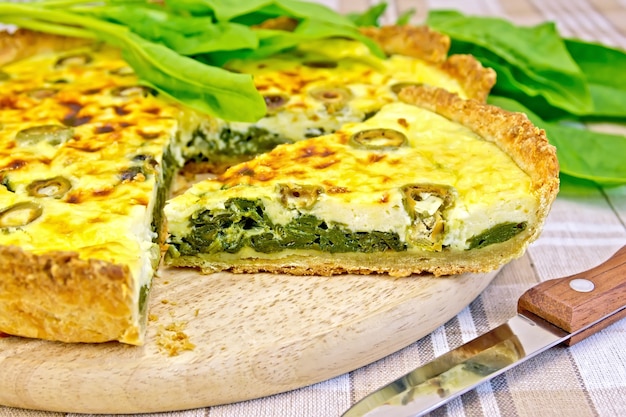 Pastel con espinacas, queso y aceitunas en una tabla redonda, hojas de espinaca, cuchillo sobre manteles de lino de fondo