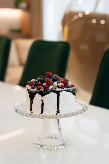 Pastel gourmet con cobertura de chocolate y bayas en un soporte de vidrio sobre una mesa blanca en un interior moderno. Enfoque selectivo suave.