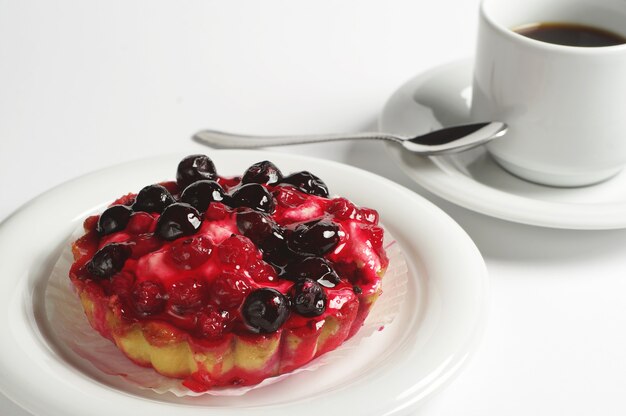 Pastel con grosellas en plato y taza de café sobre fondo blanco.