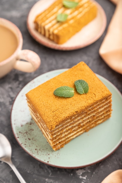 Pastel de miel casero con crema de leche y menta con una taza de café sobre una superficie de hormigón negro y textil naranja