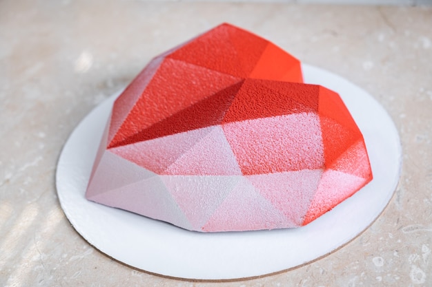 Pastel de mousse de corazón rojo facetado con revestimiento de terciopelo sobre una mesa de mármol rosa suave.