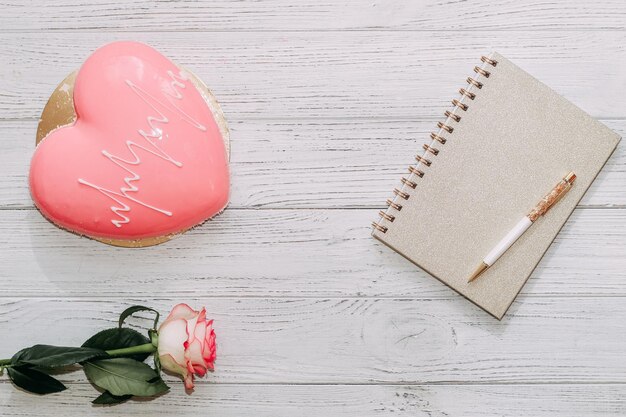 Pastel de mousse rosa en forma de corazón en la mesa con rosa y bloc de notas