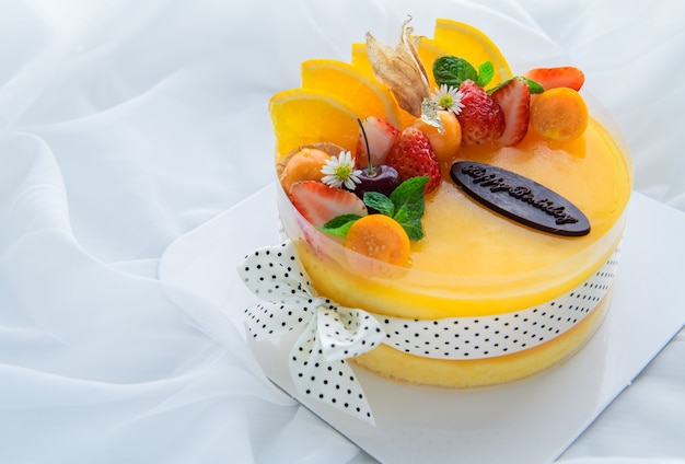 Pastel de naranja con feliz día de nacimiento y cubierto con naranja, fresa, arándano y uchuva sobre fondo de tela blanca, espacio de copia y concepto de postre