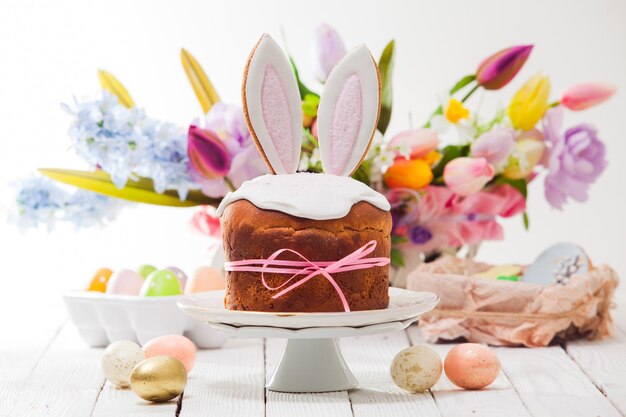 El pastel de Pascua con orejas de conejo comestibles se encuentra en un soporte Huevos pintados alrededor del pastel En el fondo ramos de coloridas flores de primavera