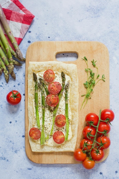 Pastel con queso crema de espárragos verdes y tomates en una tabla de cortar de madera sobre un fondo azul claro