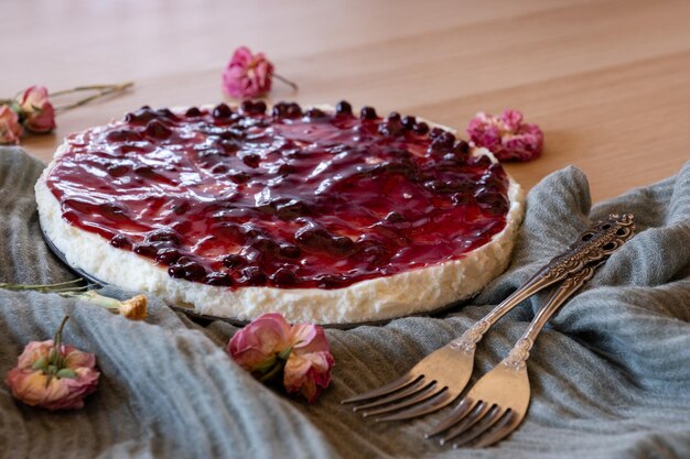 Foto pastel de queso frío con jalea de cereza y rosas a su alrededor servido sobre fondo de madera con lanza verde y tenedores antiguos