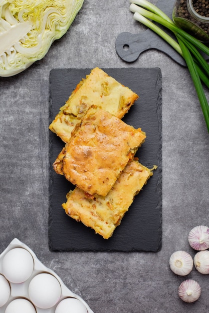 Pastel de repollo con zanahorias sobre un hormigón oscuro con ingredientes para cocinar, huevos, cebollas verdes, verduras y especias.