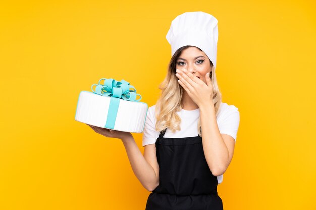 Pastelaria de menina adolescente segurando um bolo grande isolado na parede amarela com expressão facial de surpresa