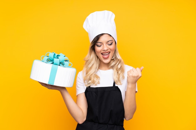 Pastelaria de menina adolescente segurando um bolo grande na parede amarela comemorando uma vitória