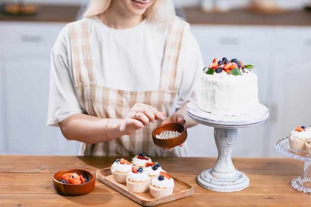 Pasteleiro confeiteiro jovem mulher caucasiana decora bolo de doces bolas doces na mesa da cozinha Bolos cupcakes e sobremesa doce