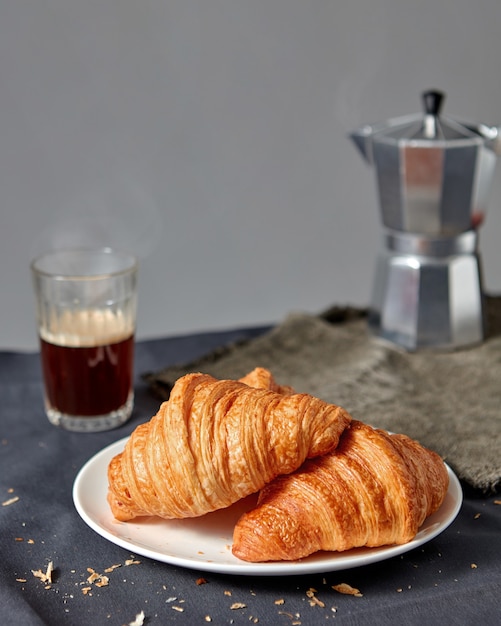 Pastelería francesa recién horneada en un plato con un vaso de café aromático caliente y géiser sobre un fondo oscuro texile, copie el espacio.